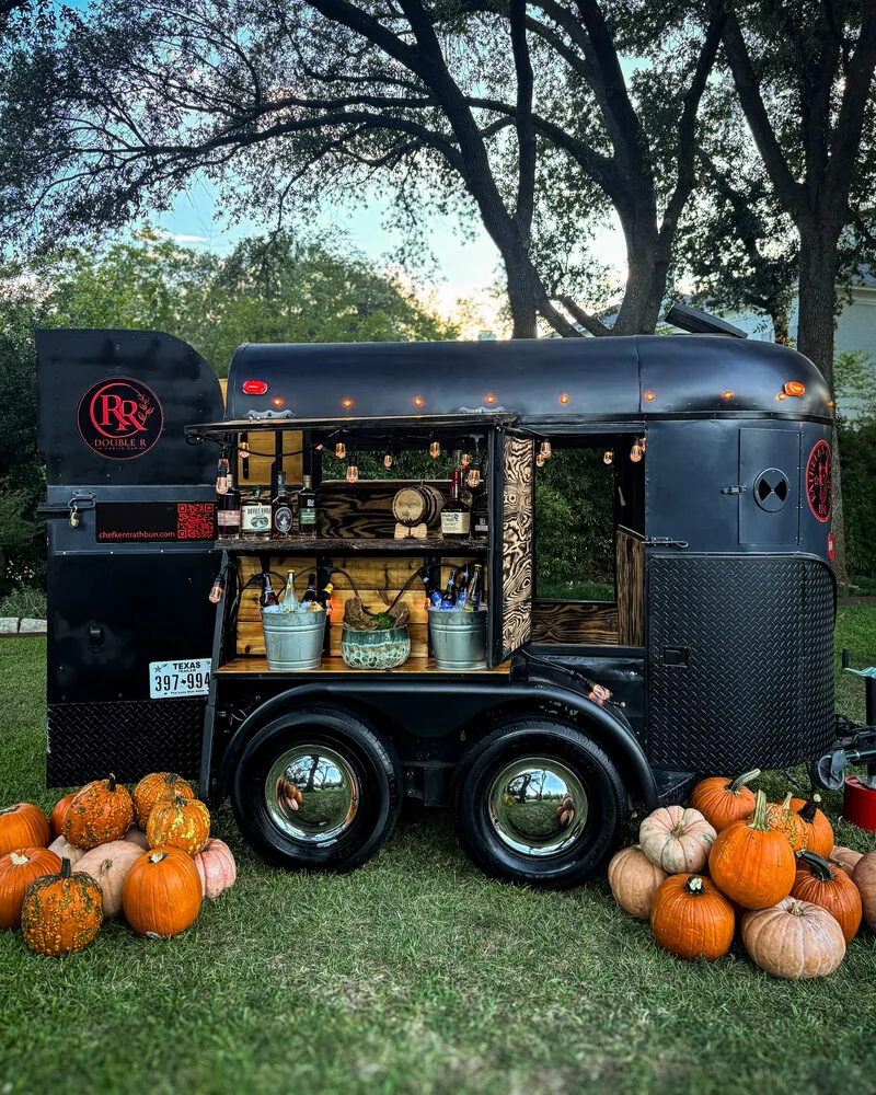 Double R's black vintage horse trailer bar, open with bottles and lanterns, surrounded by pumpkins on a lawn at dusk