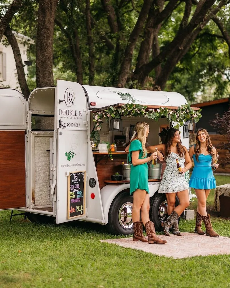 Three guests in sundresses and cowboy boots holding drinks at the Double R white vintage horse trailer bar under shade trees